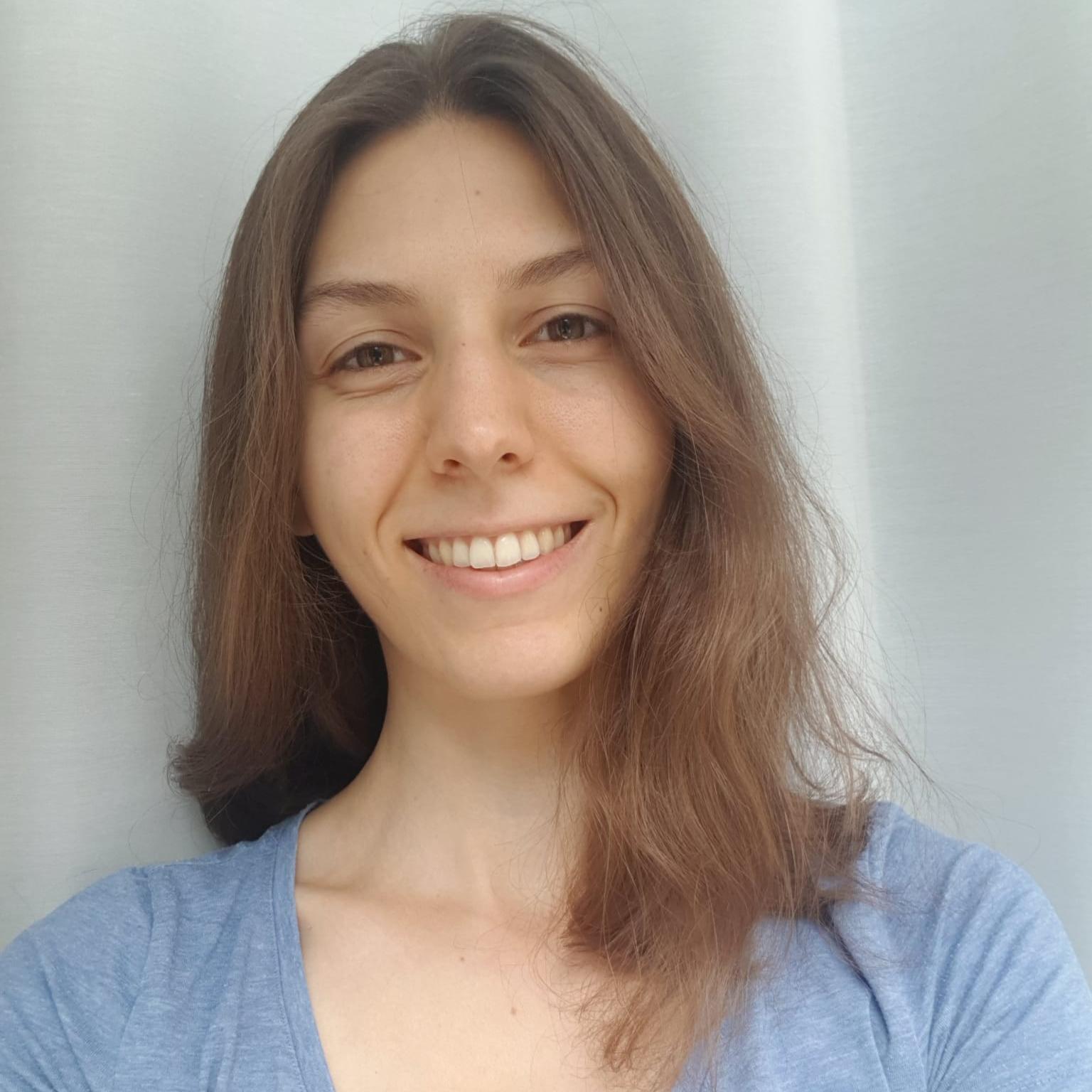 MedAT-Erfahrungsbericht Ida: Junge Frau mit mittellangen braunen Haaren, die lächelnd vor einem weißen Hintergrund steht. Sie trägt ein schlichtes, blaues Oberteil und wirkt natürlich und sympathisch.
