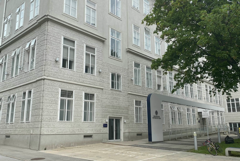 Das Gebäude der Medizinischen Universität Wien mit einer verzierten Fassade und einem modernen Eingangsbogen. Es spiegelt die Kombination von historischer Architektur und zeitgemäßen Elementen wider, die für die Universität typisch ist. Ein Fahrrad ist an einem Baum vor dem Gebäude abgestellt.