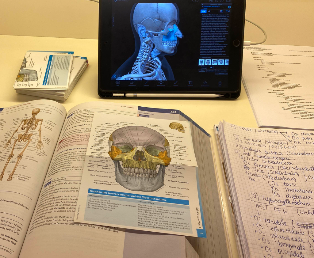 Medizinische Lernunterlagen mit einem offenen Anatomiebuch, Lernkarten, handschriftlichen Notizen und einem Tablet mit einer 3D-Anatomie-App. Das Bild zeigt den Lernprozess eines Medizinstudenten, der verschiedene Hilfsmittel für ein effektives Studium der Anatomie nutzt.