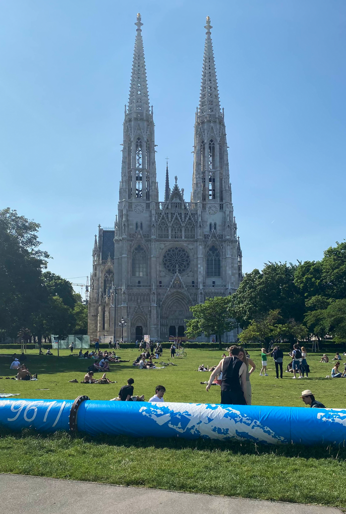 Der Votivpark in Wien mit der beeindruckenden Votivkirche im Hintergrund. Menschen sitzen und entspannen auf der grünen Wiese an einem sonnigen Tag. Im Vordergrund ist ein blauer Wasserrohrabschnitt zu sehen, der die historische Architektur der Kirche in einen modernen Kontext setzt.