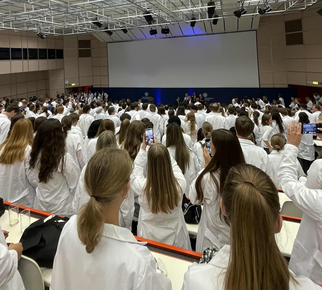 Eine White Coat Ceremony an der Medizinischen Universität Wien, bei der zahlreiche Medizinstudierende in weißen Kitteln gemeinsam einen feierlichen Moment erleben. Die Teilnehmer stehen vor einer Bühne und halten diesen besonderen Moment mit ihren Smartphones fest. Ein wichtiger Meilenstein für angehende Mediziner in Österreich.