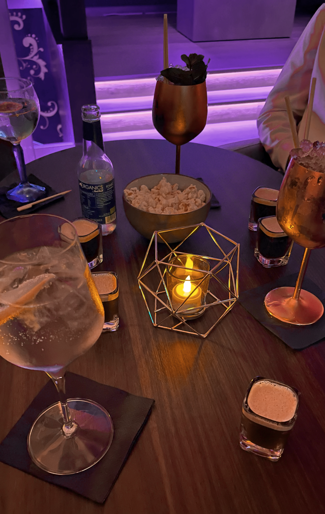 Ein Tisch in einer Bar mit verschiedenen Cocktails, Shots, einer Schale Popcorn und einer brennenden Kerze in einem dekorativen Halter. Die stimmungsvolle Beleuchtung unterstreicht das entspannte und elegante Ambiente, ideal für einen Abend in der Wiener Bar-Szene.