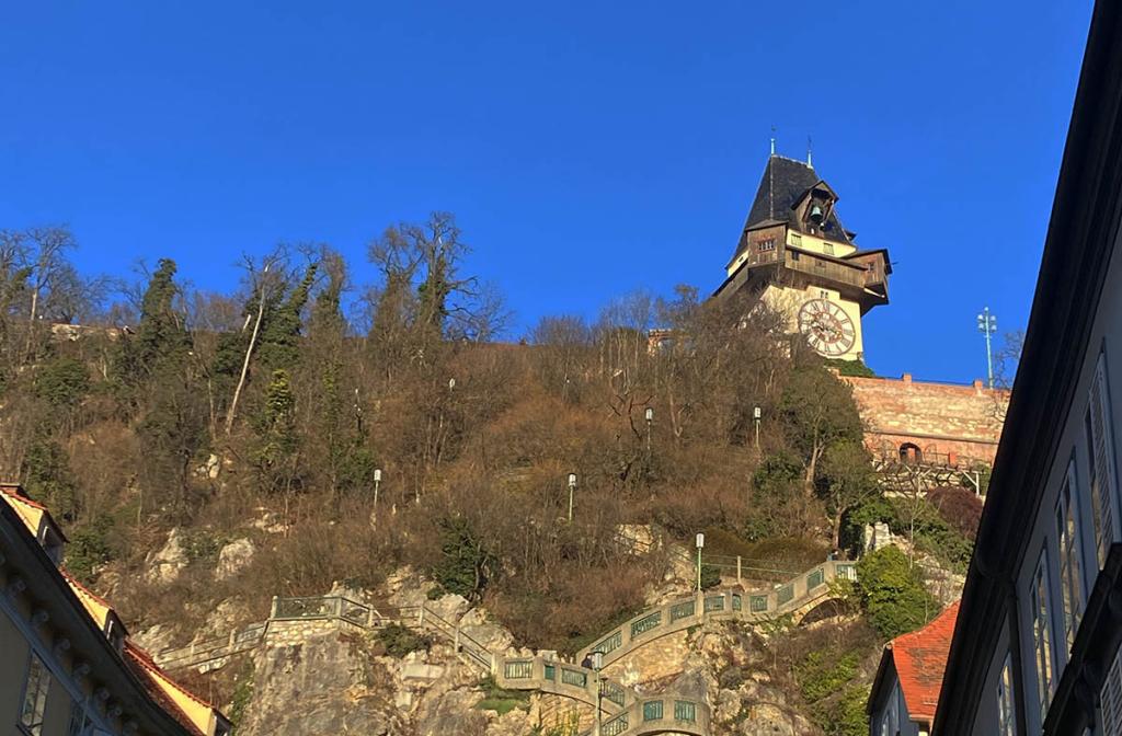 Grazer Uhrturm auf dem Schloßberg an einem sonnigen Tag, umgeben von Natur und historischer Treppe.