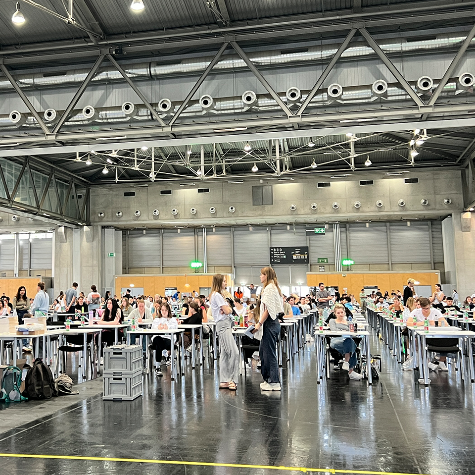 MedAT-Erfahrungsbericht: Prüfungshalle mit vielen jungen Menschen, die an einer MedAT-Aufnahmeprüfung in Wien teilnehmen. Einige Betreuer in orangefarbenen T-Shirts unterstützen die Teilnehmer. Die Halle ist groß und industriell gestaltet, mit roten Säulen und Metallstrukturen an der Decke.