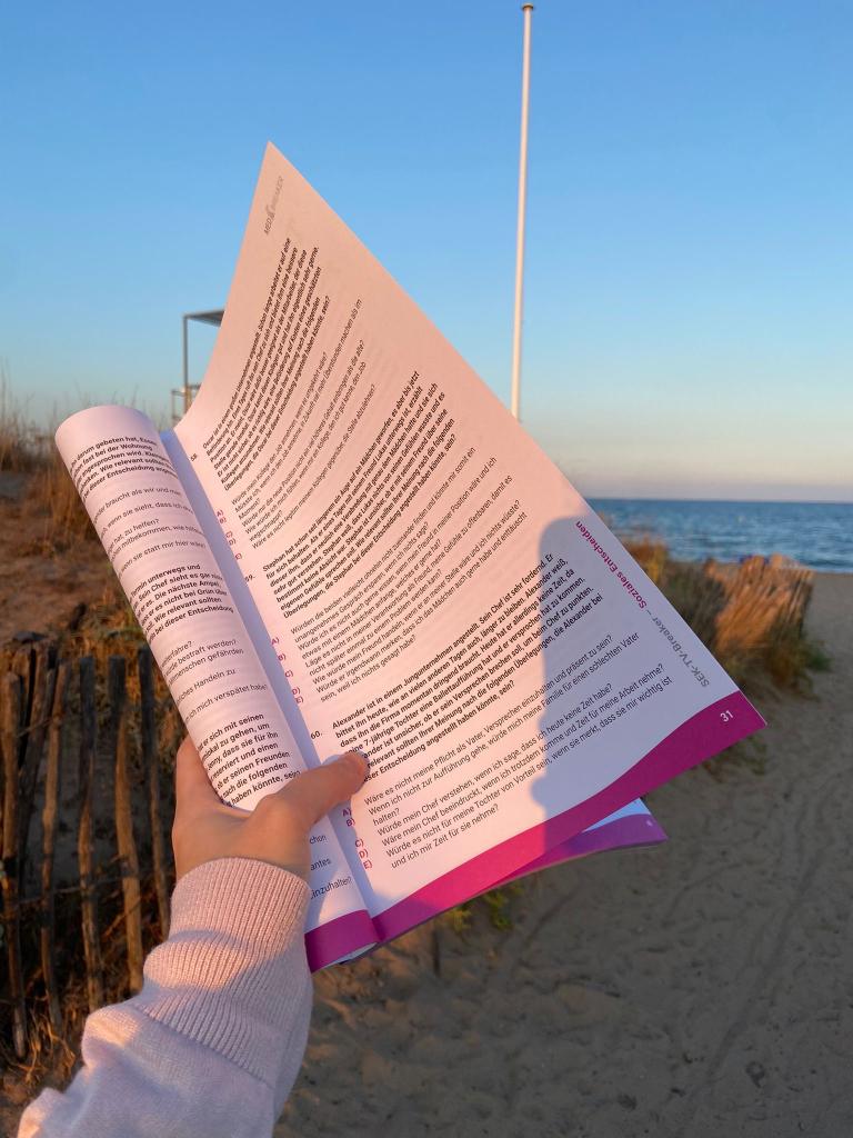 Buch statt MEDBREAKER-PDF: Person hält ein aufgeschlagenes MedAT-Übungsbuch von MEDBREAKER in der Hand, während sie am Strand steht. Im Hintergrund sind Sand, ein Holzzaun und das Meer bei Sonnenuntergang zu sehen.