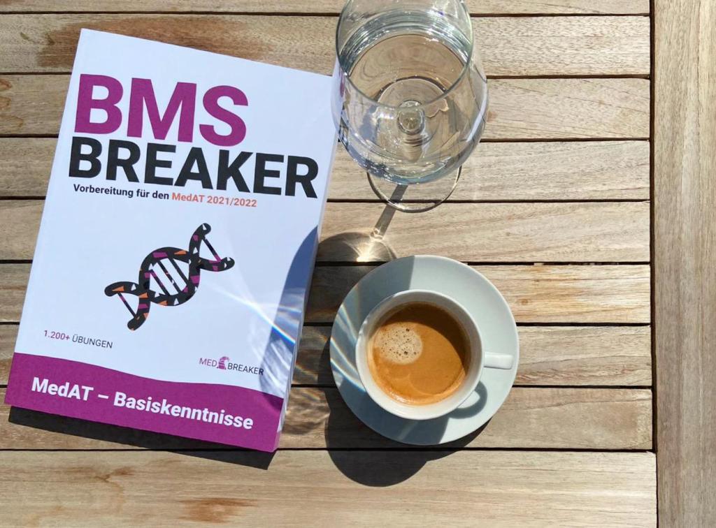Vorbereitungsbuch für den MedAT von MEDBREAKER liegt auf einem Holztisch neben einer Tasse Espresso und einem Glas Wasser – Lernpause in der Sonne.