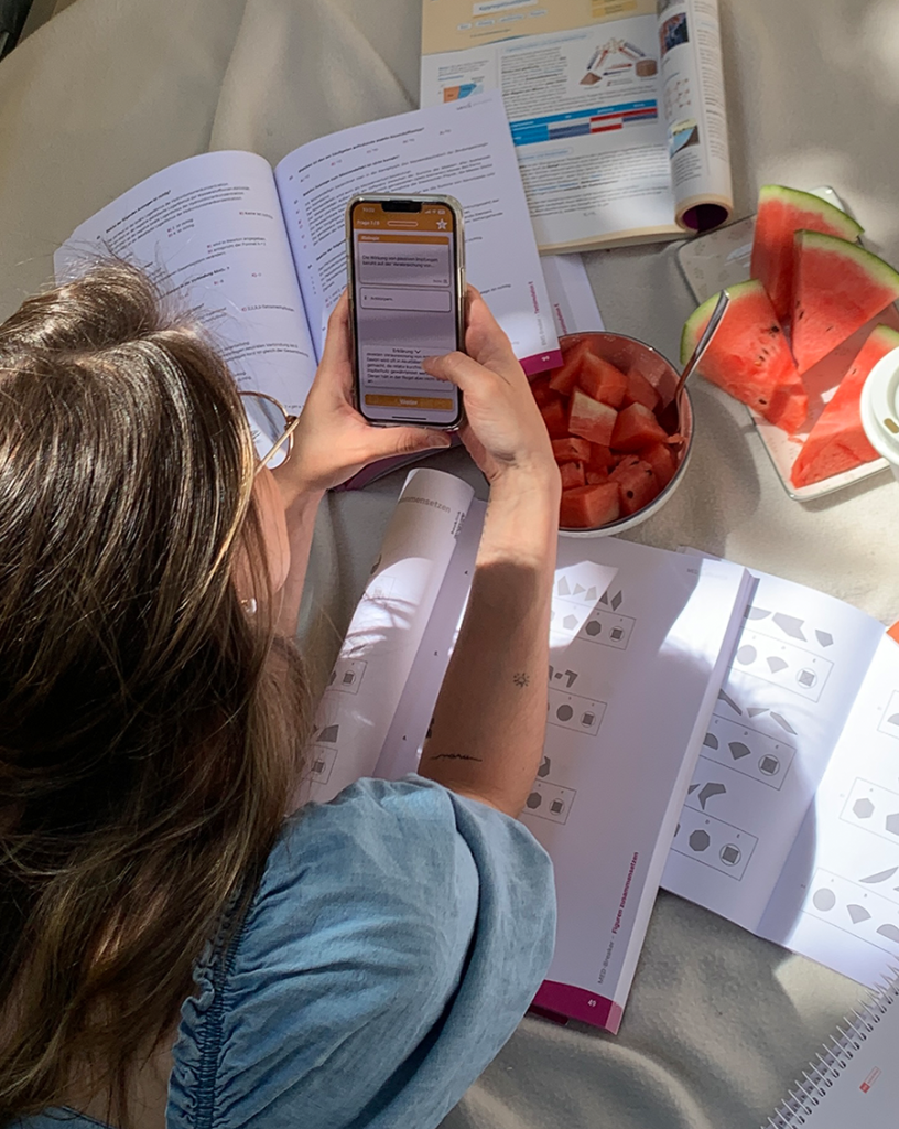 Materialien statt MEDBREAKER-PDF: Junge Frau lernt mit mehreren geöffneten Übungsbüchern für den MedAT von MEDBREAKER und nutzt ihr Smartphone die MedAT2go App, um sich Lerninhalte anzusehen. Daneben stehen ein Teller mit Wassermelone und weitere Materialien.
