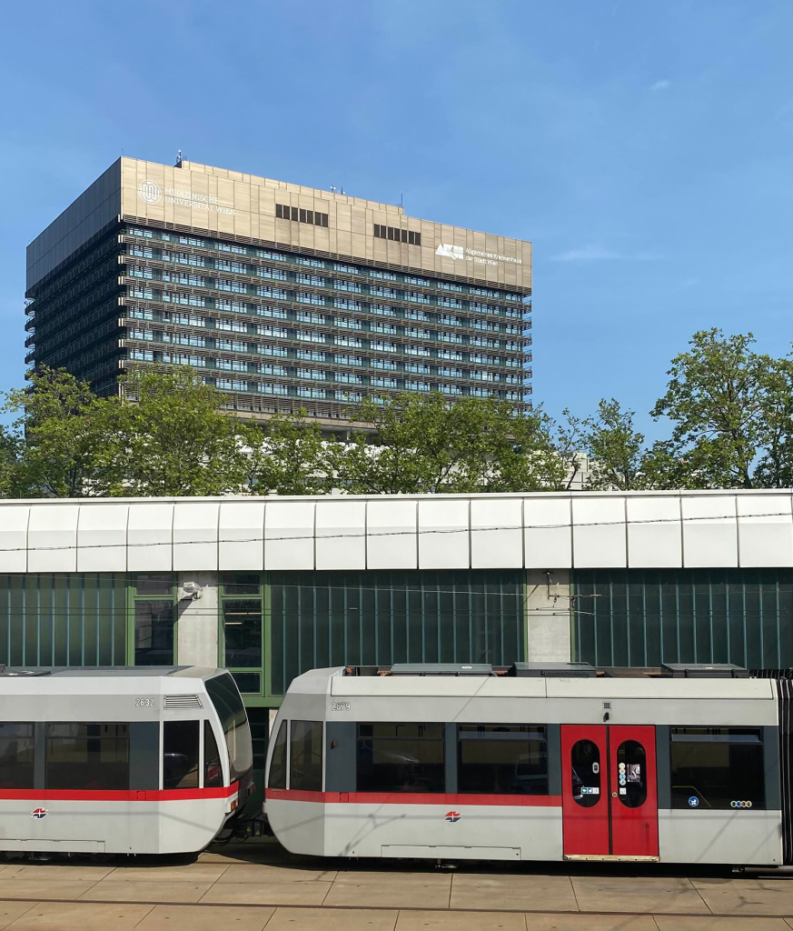 Das Gebäude des Allgemeinen Krankenhauses Wien (AKH) und der Medizinischen Universität Wien, im Vordergrund eine vorbeifahrende Wiener Straßenbahn. Die Szene zeigt die charakteristische Architektur des AKH und den öffentlichen Nahverkehr in Wien an einem sonnigen Tag.