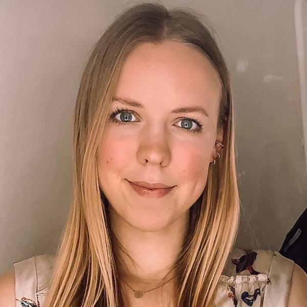 MedAT-Erfahrungsbericht Lina: Junge Frau mit langen, glatten, blonden Haaren, die freundlich in die Kamera lächelt. Sie trägt ein helles Oberteil mit floralem Muster, der Hintergrund ist neutral und weich beleuchtet.
