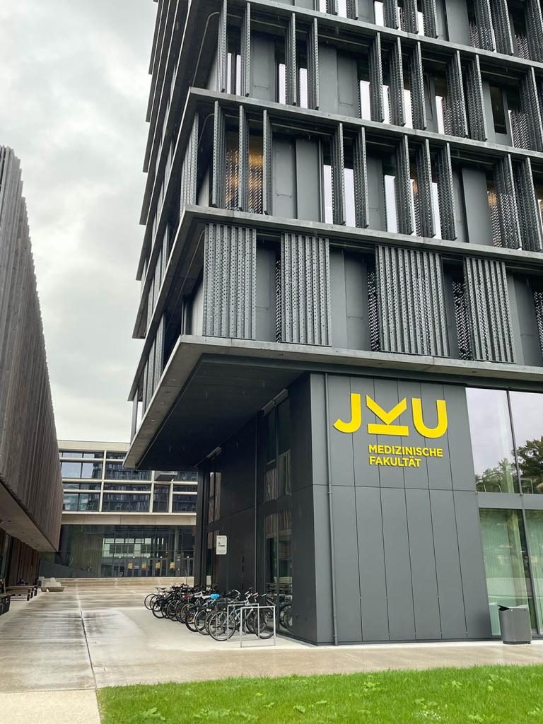 Bild der Medizinischen Fakultät der JKU Linz. Vor dem Gebäude sind Fahrräder abgestellt, während Studierende und Besucher die MedAT-Zulassung und Studienwechsel-Möglichkeiten diskutieren.