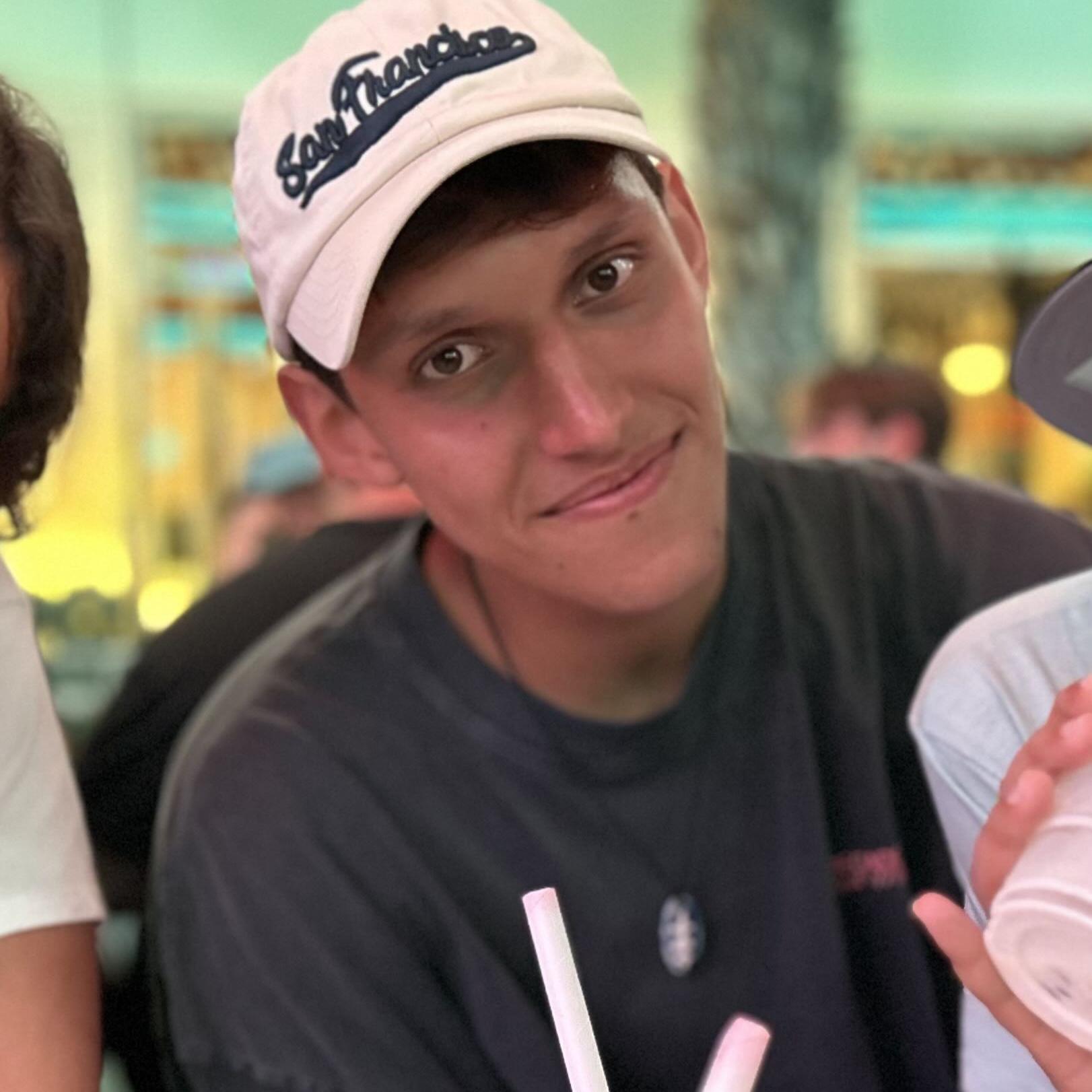 MedAT-Erfahrungsbericht Jakob: Junger Mann mit dunkler Basecap und dunklem Shirt, der an einem Tisch mit Getränken sitzt und leicht zur Kamera lächelt. Die Umgebung wirkt sommerlich und farbenfroh, vermutlich im Freien.