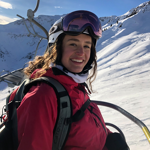 MedAT-Erfahrungsbericht Violetta: Junge Frau mit lockigem braunem Haar in Skibekleidung auf einem Sessellift. Sie trägt einen roten Anorak, Skihelm und Skibrille, im Hintergrund schneebedeckte Berge bei Sonnenschein.